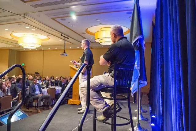 At the Bellevue Police Foundation's Annual Breakfast, two men in uniform are on stage; one stands at a podium speaking to the audience, while the other sits on a stool. Bellevue Police Foundation