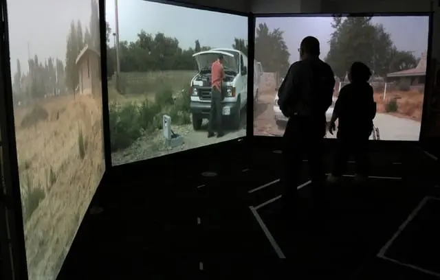 Two people stand in a room with three large screens displaying outdoor scenes, including a person near a vehicle, reminiscent of the support given by the Bellevue Police Foundation through their 2019 Grants to enhance community safety initiatives. Bellevue Police Foundation