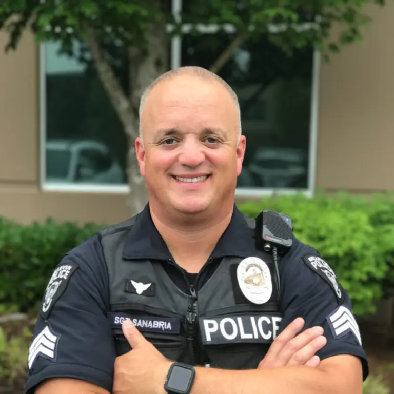 A smiling police officer in uniform stands with arms crossed in front of a building and trees, proudly representing the Bellevue Police Foundation Service Awards.