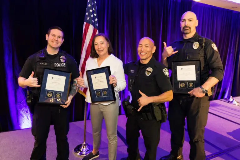 Four people in police uniforms stand on stage, proudly holding Bellevue Police Foundation Service Awards, smiling at the camera. An American flag is visible in the background.