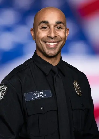 A Bellevue Police Foundation officer in uniform smiles warmly, framed by a blurred American flag in the background.