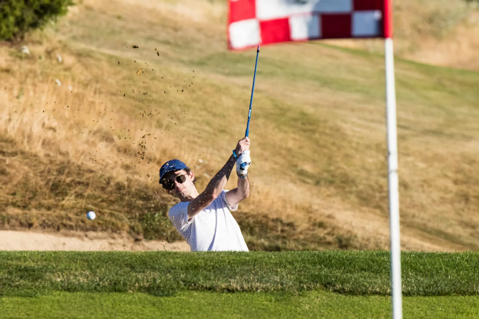 Person in white shirt hitting a golf ball from a sand bunker towards a hole marked by a red flag on a golf course.