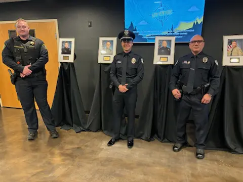 Three Bellevue Police Officers stand in front of framed photographs on draped tables, inside a room with a flat-screen display on the wall. They proudly receive the State's Highest Recognition for their exemplary service.