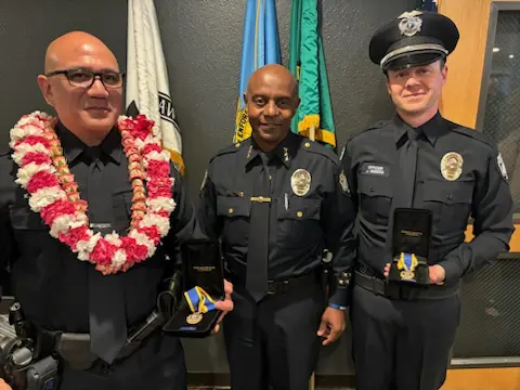 Three Bellevue Police Officers stand together; two proudly hold the State's Highest Recognition medals while the third wears a floral lei. Flags wave gently in the background, adding an air of solemn celebration to their commendation.