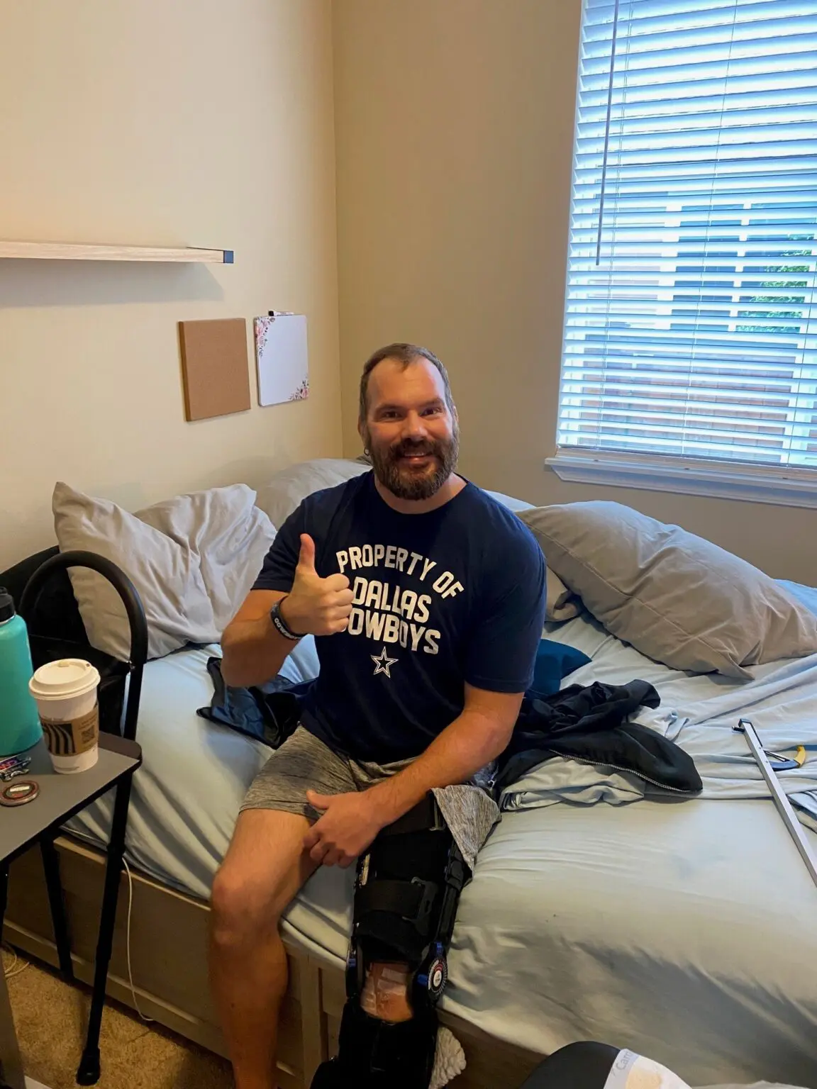 Officer Kevin Bereta sits on the bed, confidently giving a thumbs-up. Sporting a "Property of Dallas Cowboys" shirt and knee brace, his recovery update looks positive. Crutches rest against the bed while a coffee cup is on the nearby table.