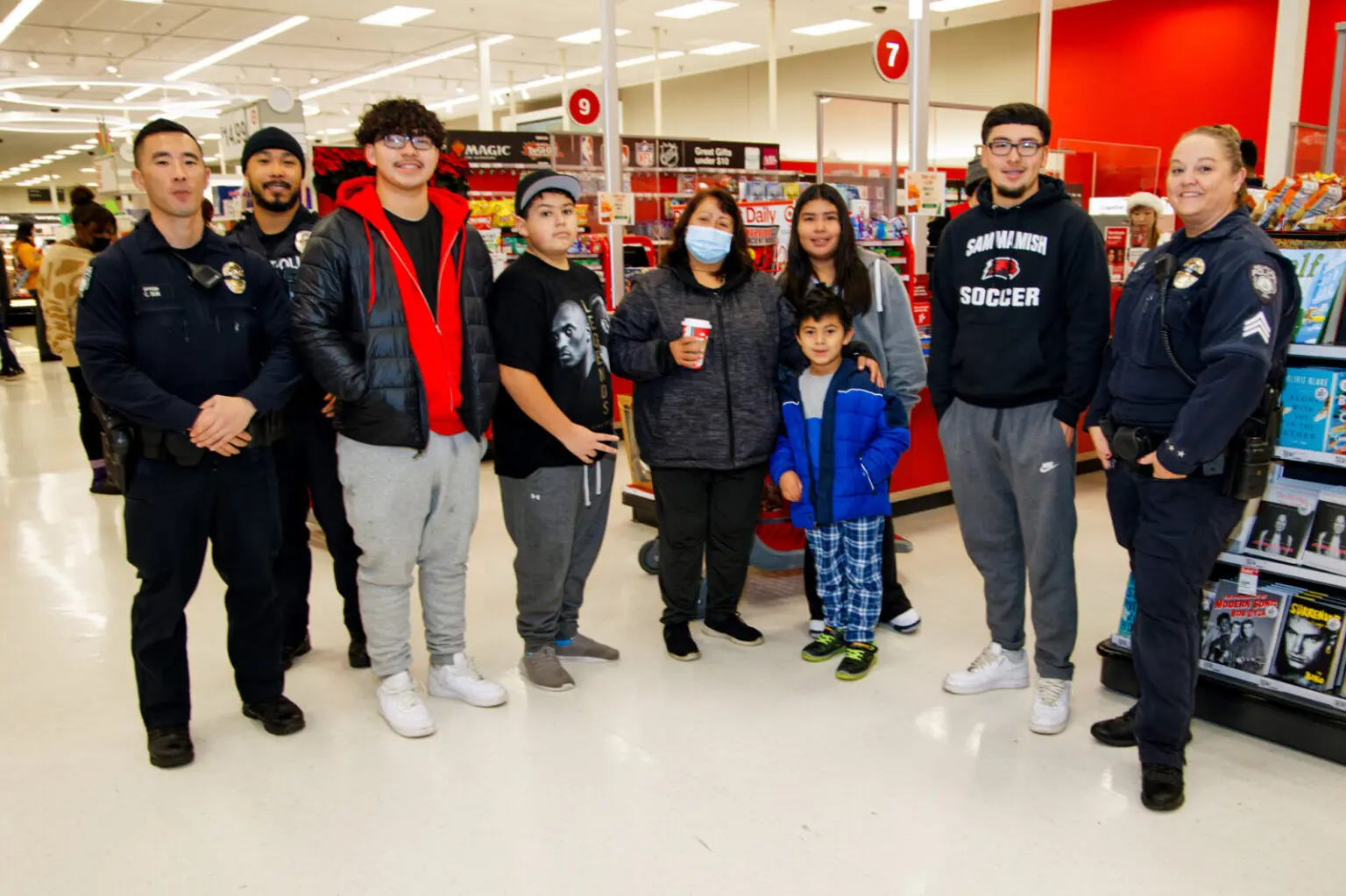 A group of four police officers and six people, including a child, share smiles while participating in the Shop with a Cop event, posing together in a store aisle.