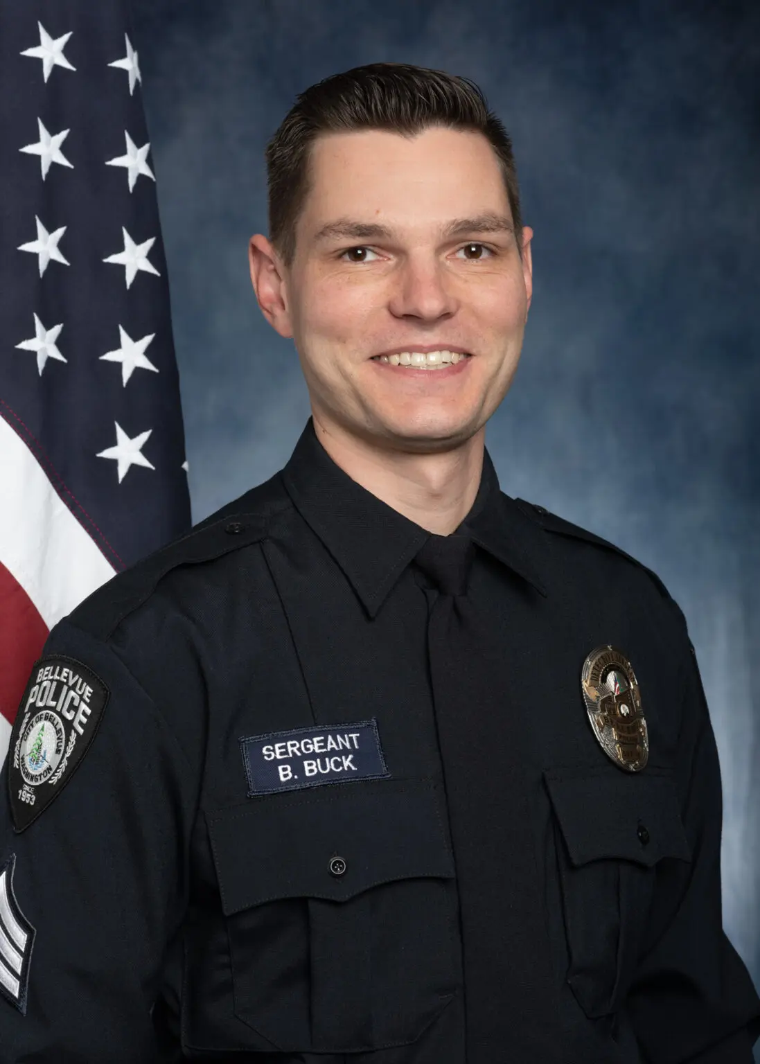 Officer Craig Hanaumi, in uniform, stands solemnly in front of an American flag, proudly displaying his 2023 Service Award.