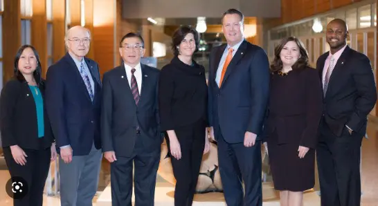 Seven people in formal attire stand in a well-lit hallway, marking a significant moment after Bellevue passes its biannual budget. Their poised stance captures a sense of accomplishment and anticipation, perhaps linked to new recruiting initiatives or upcoming foundation grants.