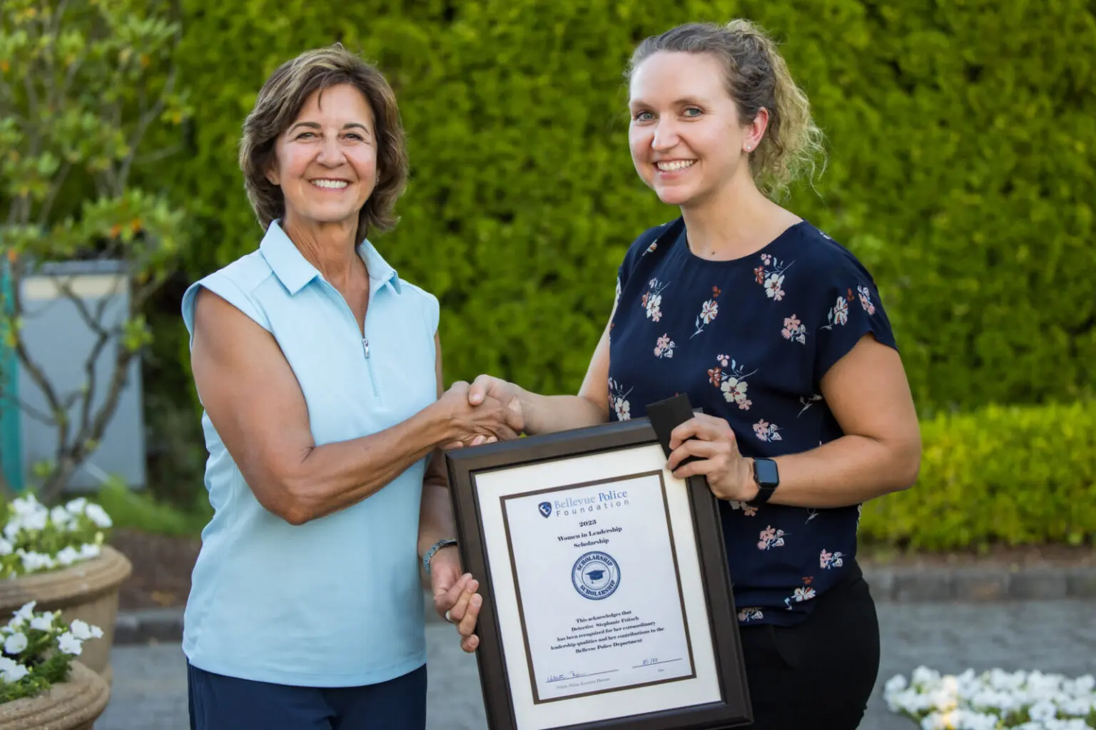 Two women smiling; one holds a framed certificate while they shake hands outside, with greenery in the background. Celebrating the success of the Women in Leadership 2023 Scholarship Program, these inspiring leaders reflect on their journey and growth.