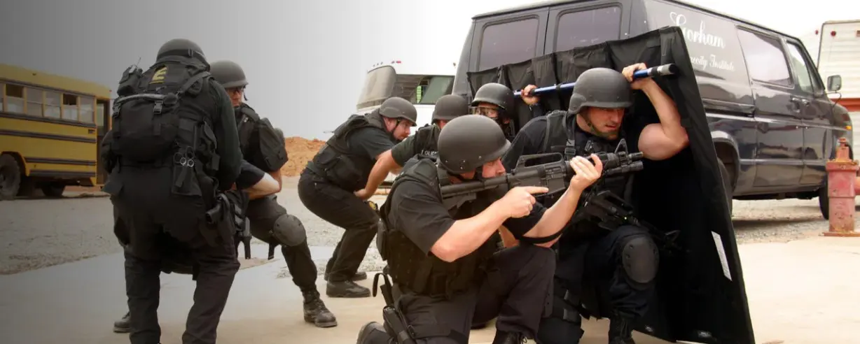 A team of armed tactical officers, equipped with public safety tools like helmets and shields, advances strategically near vehicles in an outdoor area.