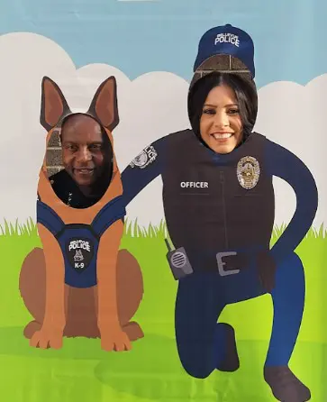 Two people pose with their faces in cutouts of a police K-9 and officer, against a cartoon background with a grassy field and clouds, celebrating National Night Out.