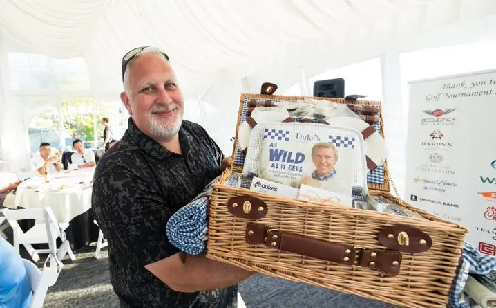 A man holding a wicker basket with a poster inside stands in a white tent at the awards banquet, adding an elegant touch to the event's atmosphere.