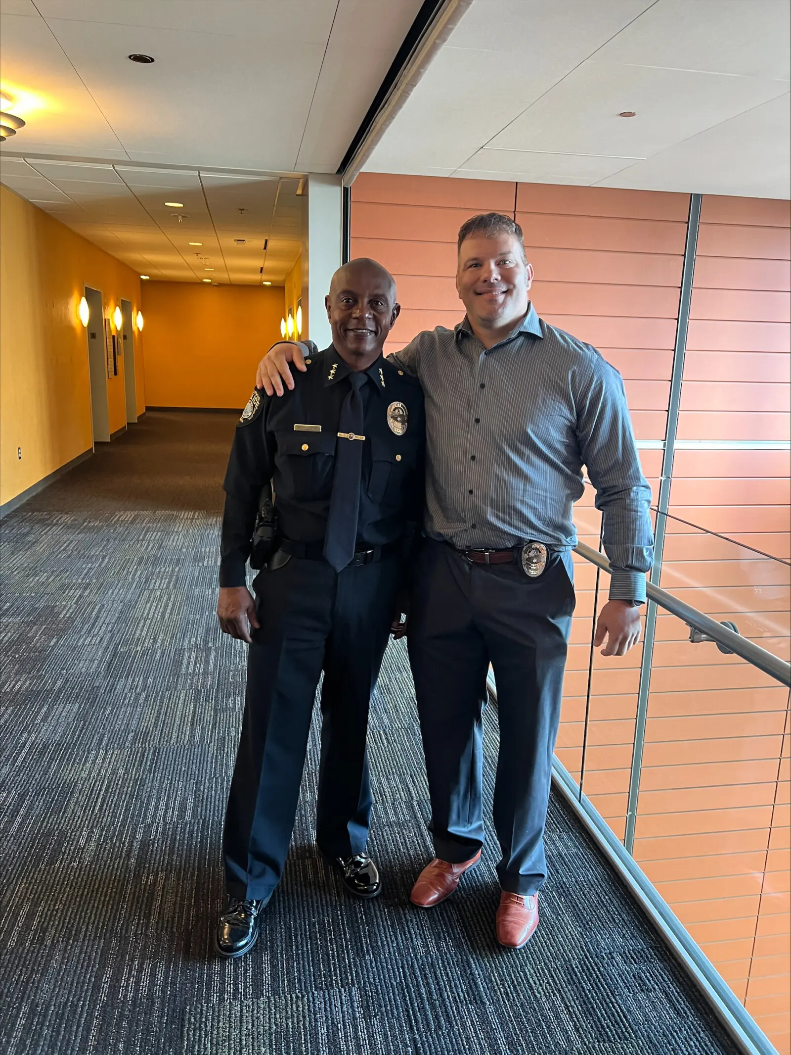 Officer Kevin Bereta stands in a hallway, proudly wearing his formal police uniform. Beside him, a man in a gray shirt with a badge clipped to his belt shares a warm smile. They are happily posing with their arms around each other.