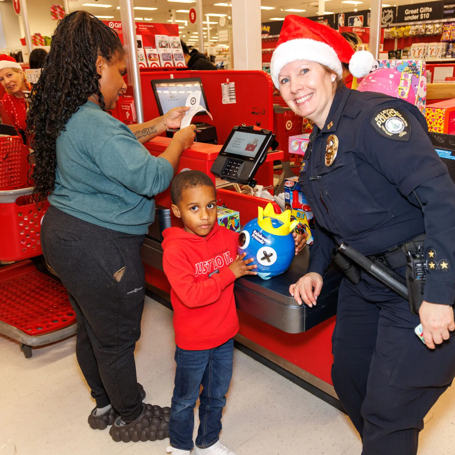 A police officer in a Santa hat, part of the Heroes and Helpers program, smiles at a boy holding a toy at the checkout counter while a woman pays during a Bellevue Police Foundation holiday event.