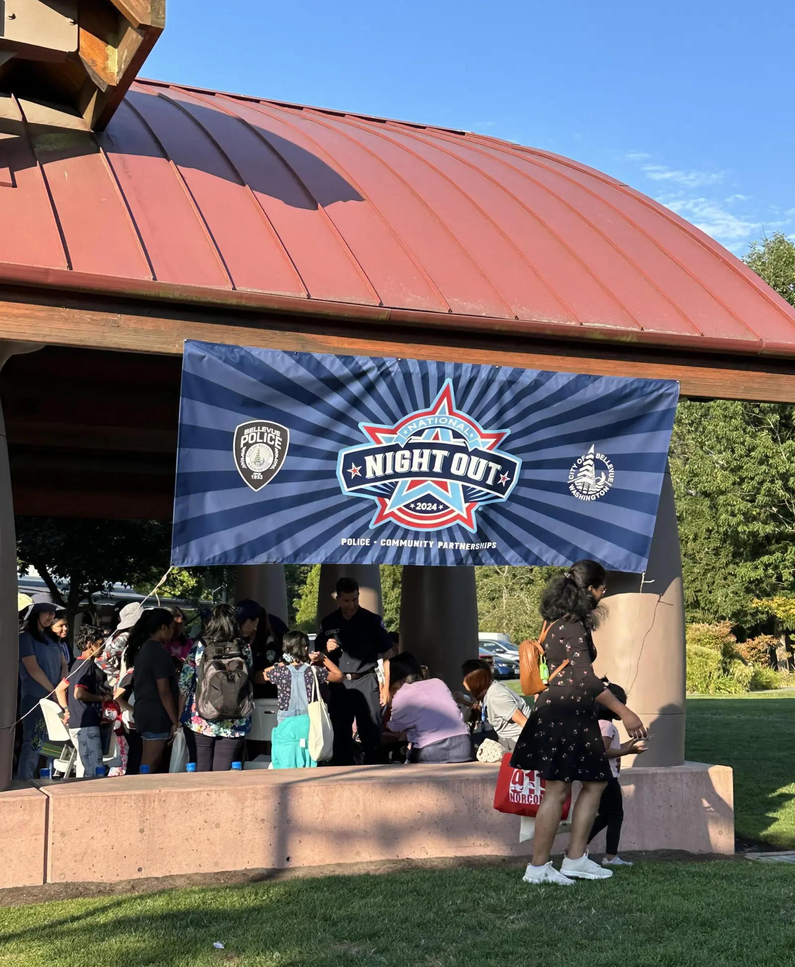 A community gathering under a shelter features a banner that reads "Night Out 2024." People stand and chat in Bellevue's park setting, celebrating National Night Out.