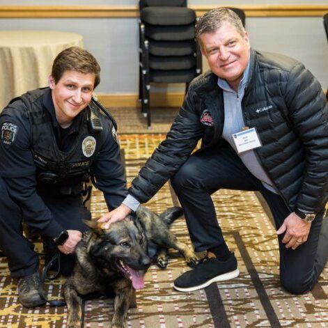 At the Bellevue Police Foundation Annual Breakfast, a police officer and a man crouch next to a German Shepherd indoors, both smiling as they pet the dog.