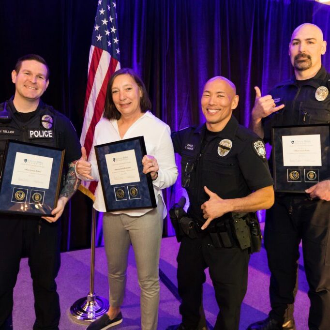 Four people in police uniforms stand on stage, proudly holding Bellevue Police Foundation Service Awards, smiling at the camera. An American flag is visible in the background.