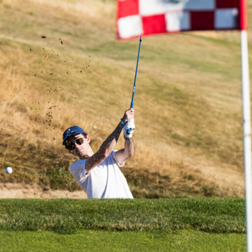 Person in white shirt hitting a golf ball from a sand bunker towards a hole marked by a red flag on a golf course.