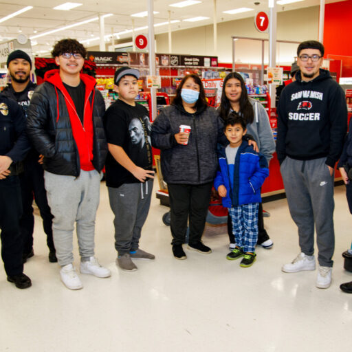 A group of four police officers and six people, including a child, share smiles while participating in the Shop with a Cop event, posing together in a store aisle.