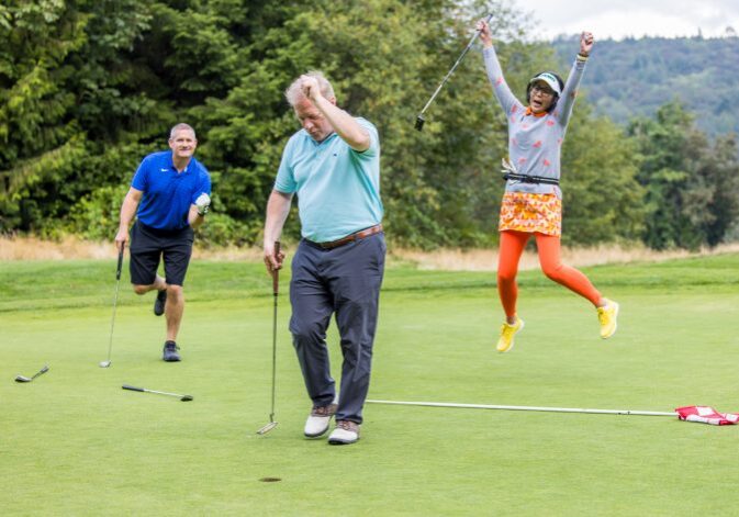 Three people enjoy a sunny day on the golf course: one man looks frustrated near a hole, another watches intently, and a woman joyfully jumps in the air with her golf club. It's just another exciting moment at one of the Bellevue Police Foundation Events.