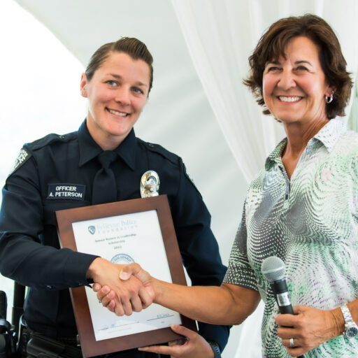 At the Bellevue Police Foundation banquet, an officer in uniform receives an award plaque and shakes hands with a smiling woman holding a microphone.