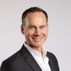 Smiling man in a suit with a white shirt against a plain background.