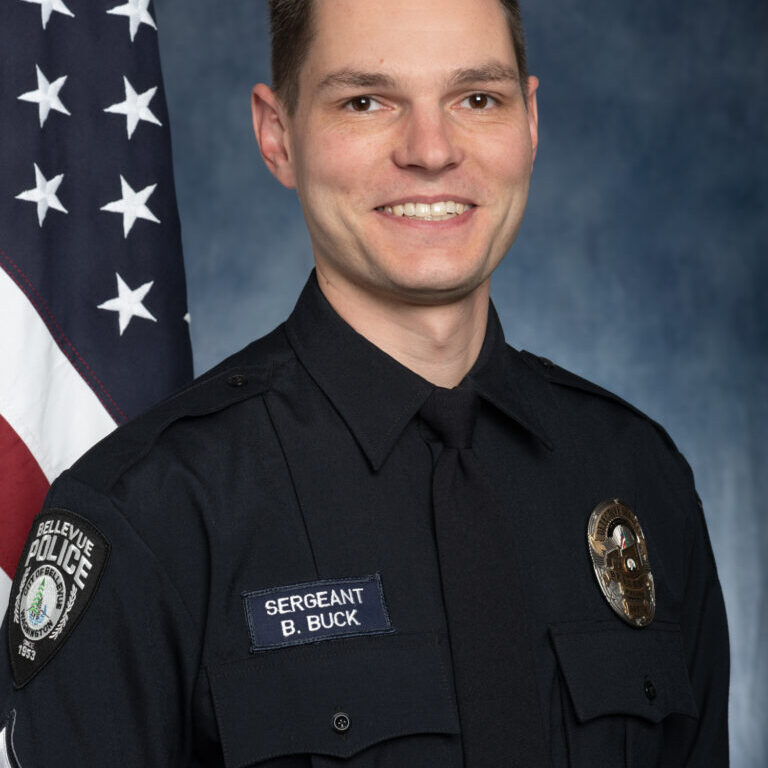 Officer Craig Hanaumi, in uniform, stands solemnly in front of an American flag, proudly displaying his 2023 Service Award.