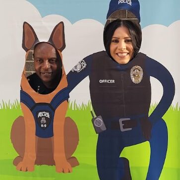 Two people pose with their faces in cutouts of a police K-9 and officer, against a cartoon background with a grassy field and clouds, celebrating National Night Out.