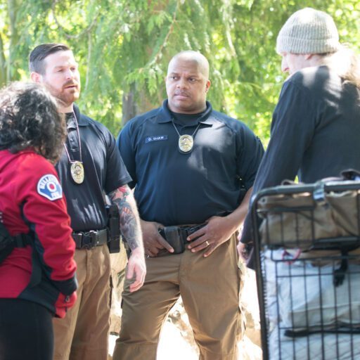 Two uniformed officers engage with a person carrying a backpack and another wearing a red jacket in a wooded area, highlighting the community outreach initiatives of Bellevue Police Foundation Programs.