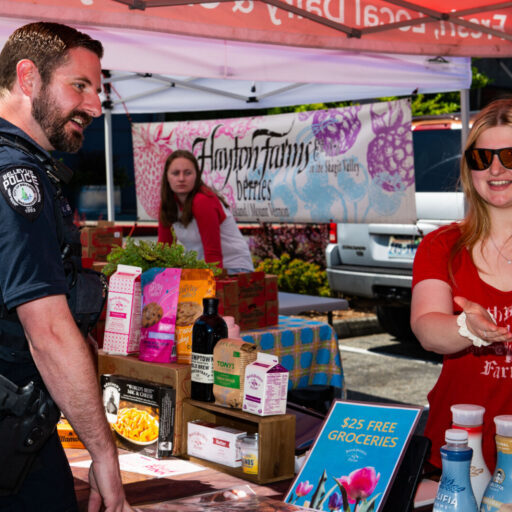 A Bellevue Police officer engages with a vendor at an outdoor market stall displaying various goods, offering foundation help through community interaction.