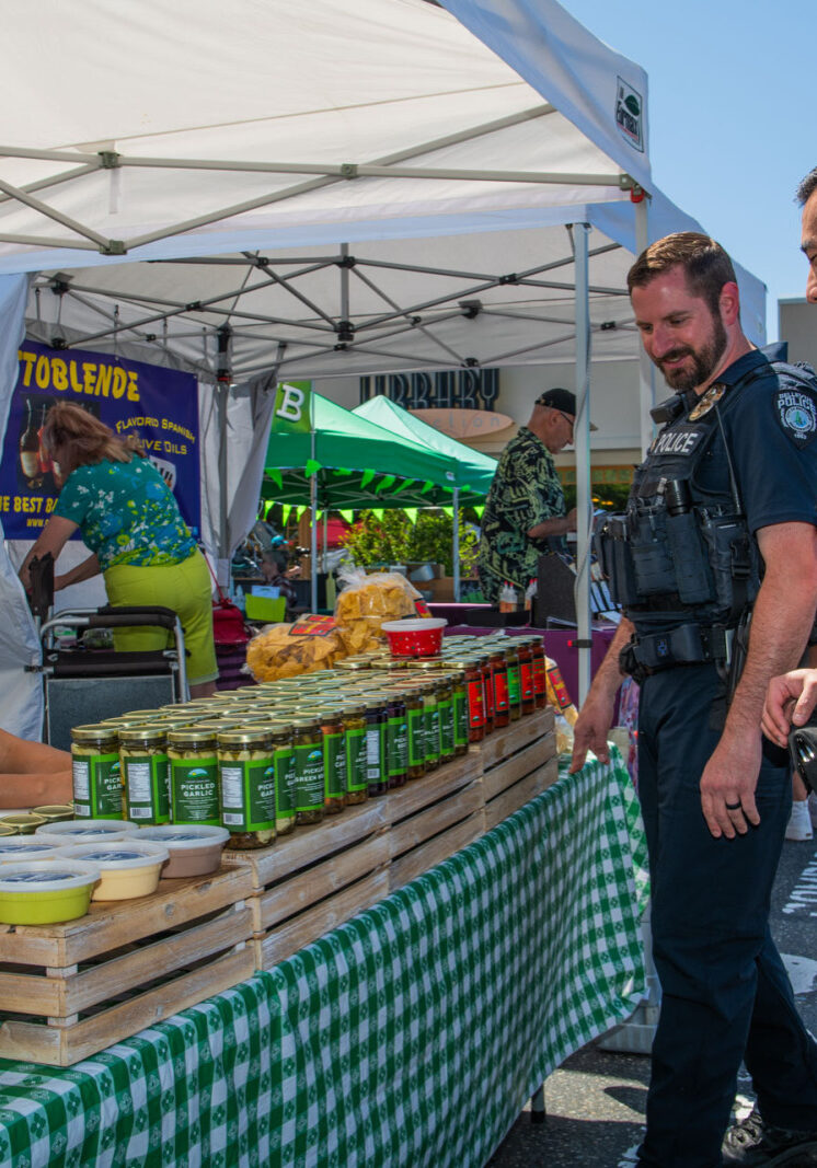 Two police officers stand at a farmers market booth, interacting with a vendor displaying various products on a checkered tablecloth. As part of an initiative supported by the Bellevue Police Foundation, they engage warmly with the community while exploring locally sourced goods.