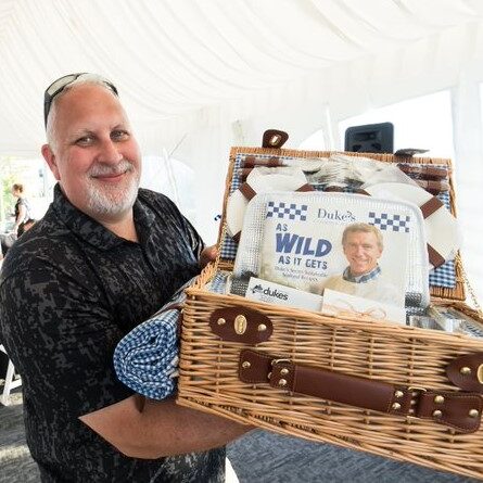 A man holding a wicker basket with a poster inside stands in a white tent at the awards banquet, adding an elegant touch to the event's atmosphere.