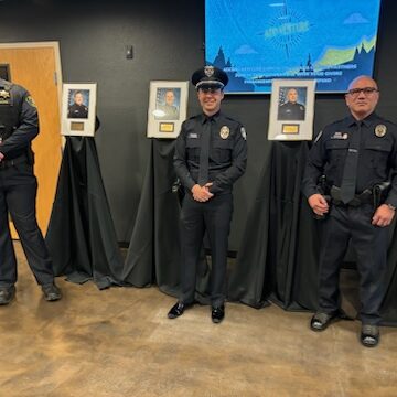 Three Bellevue Police Officers stand in front of framed photographs on draped tables, inside a room with a flat-screen display on the wall. They proudly receive the State's Highest Recognition for their exemplary service.