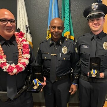 Three Bellevue Police Officers stand together; two proudly hold the State's Highest Recognition medals while the third wears a floral lei. Flags wave gently in the background, adding an air of solemn celebration to their commendation.