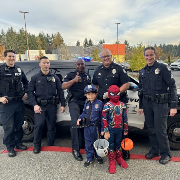 Bellevue Police officers pose with two children dressed as a police officer and Spider-Man in front of a police car, creating some Halloween fun in the parking lot.