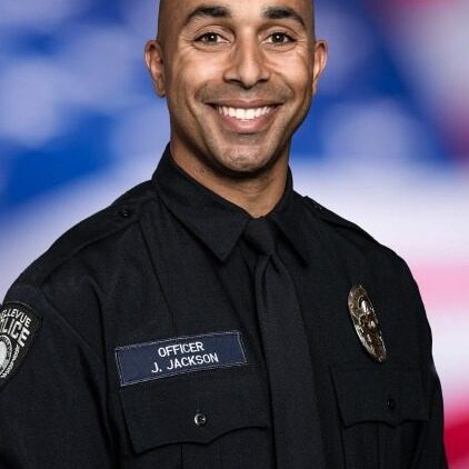 A Bellevue Police Foundation officer in uniform smiles warmly, framed by a blurred American flag in the background.