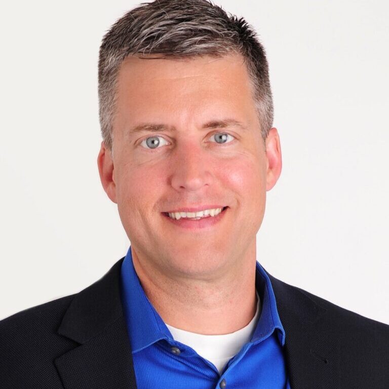 A man in a blue shirt and black blazer, a member of the Bellevue Police Foundation Board of Directors, smiles at the camera against a plain background.