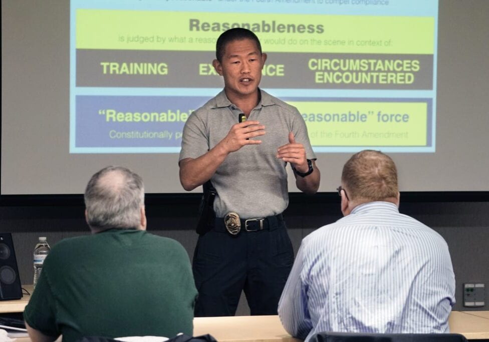 A man in a gray polo shirt conducts a presentation on reasonableness and use of force. Three people listen attentively in front of a projected slide. Bellevue Police Foundation