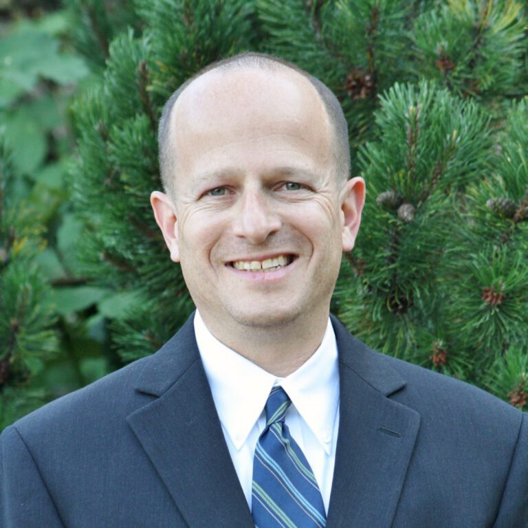 Marc Milstein, dressed in a suit and tie, smiles warmly against a backdrop of lush green foliage.