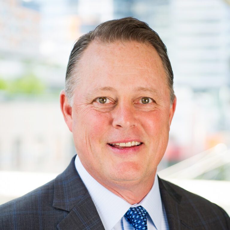 A person in a suit and blue tie, representing the Bellevue Police Foundation Board of Directors, smiles at the camera with a blurred cityscape in the background.