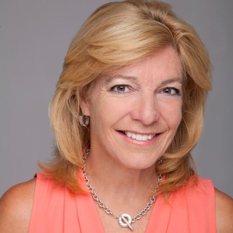 A woman with blonde hair smiles, wearing a coral top and silver necklace, against a neutral background.