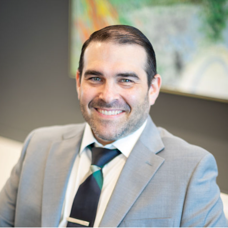 Tommy Martija, dressed in a sleek grey suit and tie, smiles confidently while seated in front of a colorful abstract painting.