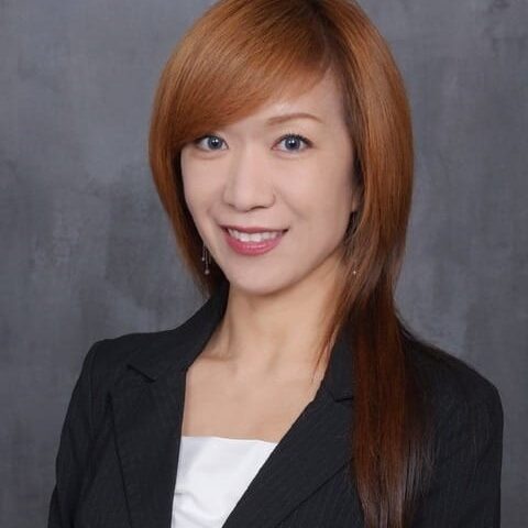 A person with long auburn hair is smiling, wearing a black blazer and a white blouse, against a gray background.