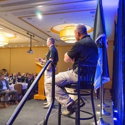 At the Bellevue Police Foundation's Annual Breakfast, two men in uniform are on stage; one stands at a podium speaking to the audience, while the other sits on a stool. Bellevue Police Foundation