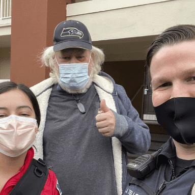 Three people wearing masks pose for a selfie, one giving a thumbs-up, representing community spirit that supports Bellevue Police Foundation Programs. A fourth person stands partially visible to the side, adding a touch of mystery to this moment captured in unity and support.