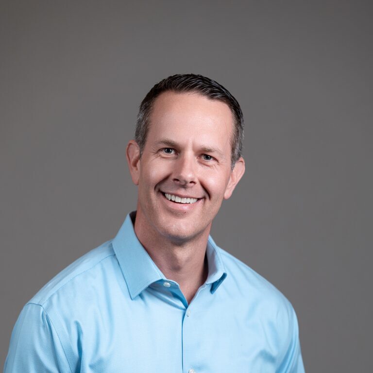 A man in a light blue shirt, a proud member of the Bellevue Police Foundation Board of Directors, smiles against a plain gray background.