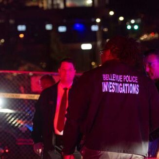 Four individuals, including someone wearing a "Bellevue Police Investigations" jacket with the Bellevue Police Foundation logo, stand near a police vehicle at night, illuminated by the flashing lights in the background.