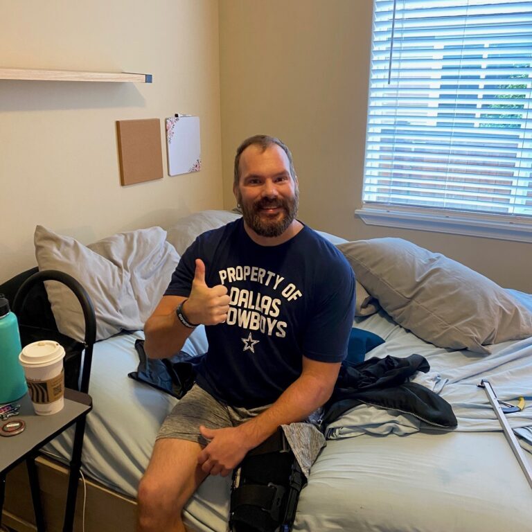 Officer Kevin Bereta sits on the bed, confidently giving a thumbs-up. Sporting a "Property of Dallas Cowboys" shirt and knee brace, his recovery update looks positive. Crutches rest against the bed while a coffee cup is on the nearby table.