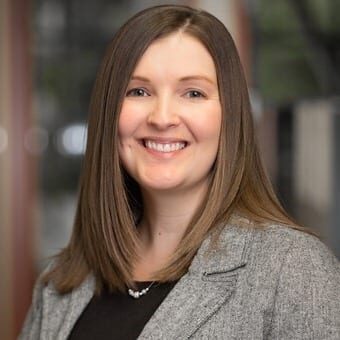 Shauna Hovancsek, with shoulder-length brown hair, smiles warmly while wearing a gray blazer over a black top, standing indoors. Bellevue Police Foundation