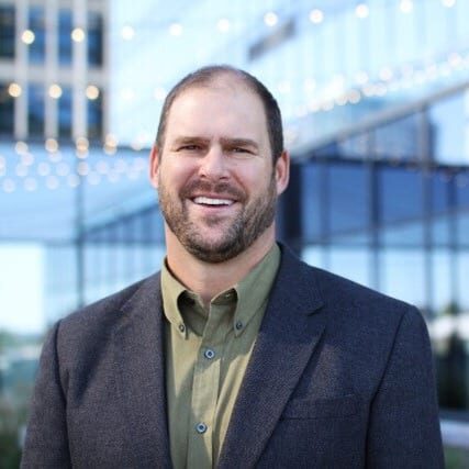 Steve Hoffmann, dressed in a suit jacket and green shirt, smiles outdoors with a modern glass building and lights in the background. Bellevue Police Foundation